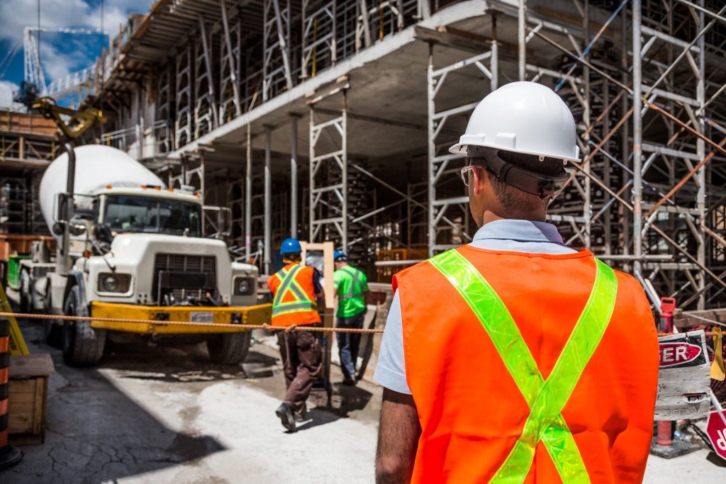 A construction site with a worker in the foreground wearing an orange safety vest and white hard hat. In the background, other accredited workers in safety gear walk near a cement mixer truck, with scaffolding and an unfinished building structure visible. This team is part of Constructionline's trusted network.