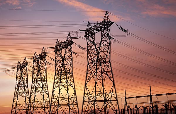A row of electricity pylons and power lines set against a vibrant sunset sky, with hues of orange, pink, and purple, forms a stunning homepage backdrop. The structures stand tall, carrying cables across the landscape. In the background, additional infrastructure is visible under the colorful sky.