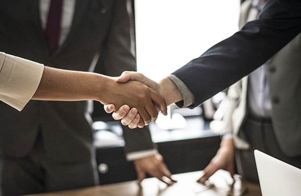 Two people in formal business attire shaking hands, suggesting a professional agreement or partnership. Both individuals are standing, with additional blurred figures and office furniture visible in the background—a snapshot perfect for any homepage or main page on a corporate website.