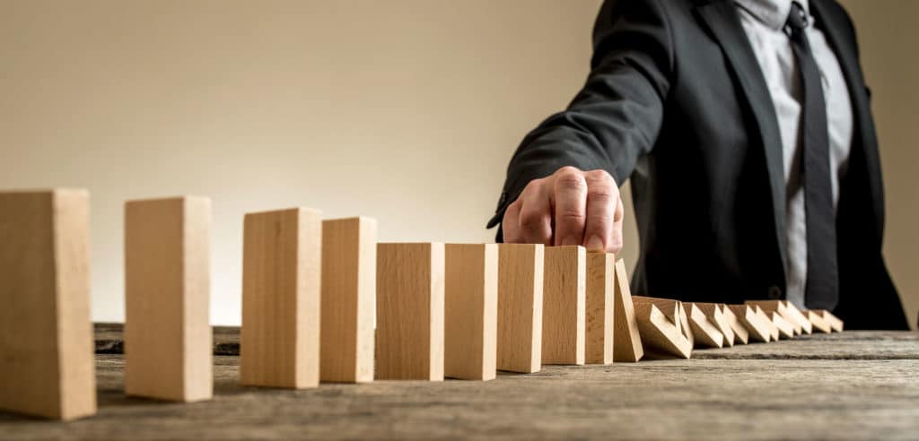 A person in a suit reaches out to stop a row of falling wooden dominoes on a wooden table. The image illustrates the concept of risk management, emphasizing its importance in preventing a chain reaction or taking action to mitigate a cascade of events.