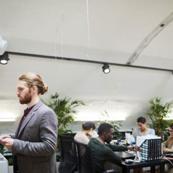 An office setting with a man in a gray blazer standing and using a copier or printer. In the background, several colleagues of diverse backgrounds are sitting at desks, working on computers, collaborating, and ensuring audit readiness. The office has plants and modern decor.