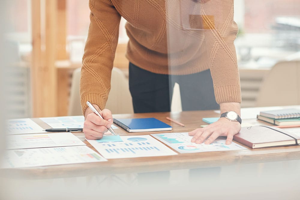 A person in a brown sweater leans over a desk, analyzing and writing on documents with charts and graphs. A notebook, additional papers, and a laptop are also visible on the desk. The scene is set in a bright workspace with large windows, highlighting efforts to grow your business during the coronavirus pandemic.