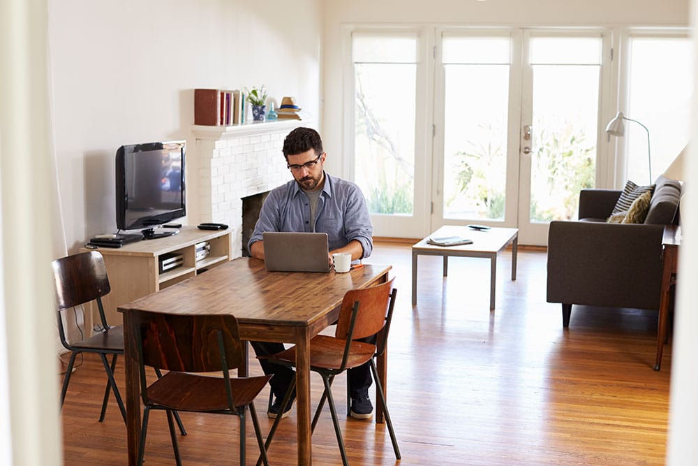 A person with glasses is sitting at a wooden dining table in a bright living room, conducting a remote gap analysis on their laptop. The room has a TV, chairs, a couch, a coffee table, and a large window providing natural light.