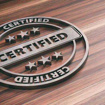 Circular metal emblem with the word "CERTIFIED" in bold letters displayed twice, one horizontally across the middle and once around the outer edge. Three stars are placed above and below the horizontal text. The emblem, signifying Achilles certification, is laid on a polished wood surface.