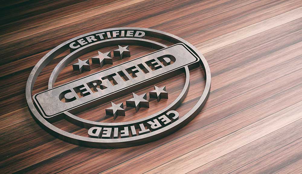 Circular metal emblem with the word "CERTIFIED" in bold letters displayed twice, one horizontally across the middle and once around the outer edge. Three stars are placed above and below the horizontal text. The emblem, signifying Achilles certification, is laid on a polished wood surface.