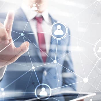 A person in a suit and tie is interacting with a virtual interface by touching one of several interconnected icons representing people. The person holds a tablet in their other hand, appearing in a blurred background with a network of lines and nodes, highlighting the importance of Achilles UVDB qualification.