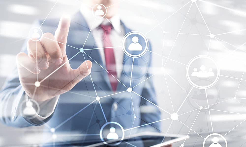 A person in a suit and tie is interacting with a virtual interface by touching one of several interconnected icons representing people. The person holds a tablet in their other hand, appearing in a blurred background with a network of lines and nodes, highlighting the importance of Achilles UVDB qualification.