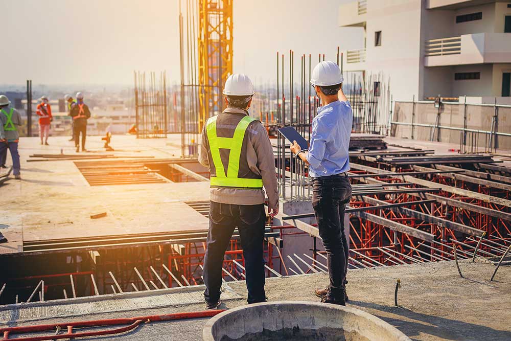 Two construction workers wearing safety vests and helmets stand on a building site, building confidence as they discuss plans on a tablet. Metal rods and scaffolding are visible, with other workers in the background. The sun is setting, casting a warm glow over the accredited project.
