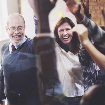 A group of people, including an elderly man with glasses and a beard, and a woman with long hair, are standing indoors and raising their hands in the air while smiling and laughing, appearing to celebrate or cheer together after achieving Full Marks in their latest Achilles UVDB Audit.
