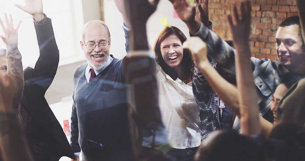 A group of people, including an elderly man with glasses and a beard, and a woman with long hair, are standing indoors and raising their hands in the air while smiling and laughing, appearing to celebrate or cheer together after achieving Full Marks in their latest Achilles UVDB Audit.