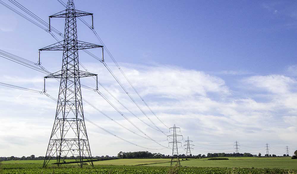 A series of tall electrical transmission towers stand in a field with green grass and crops, under a clear, blue sky. Power lines stretch between the towers, extending into the distance. Trees are visible on the horizon, symbolizing sustainable progress through reliable infrastructure and Achilles UVDB registration service.