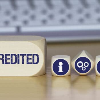 Five wooden blocks sit on a desk in front of a keyboard. The large block on the left reads "ACCREDITED," while the smaller blocks display icons: an information symbol, gears, an exclamation mark, and a thumbs-up. These symbols represent various aspects of life and the benefits accreditation brings.