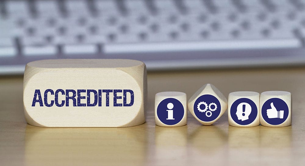 Five wooden blocks sit on a desk in front of a keyboard. The large block on the left reads "ACCREDITED," while the smaller blocks display icons: an information symbol, gears, an exclamation mark, and a thumbs-up. These symbols represent various aspects of life and the benefits accreditation brings.