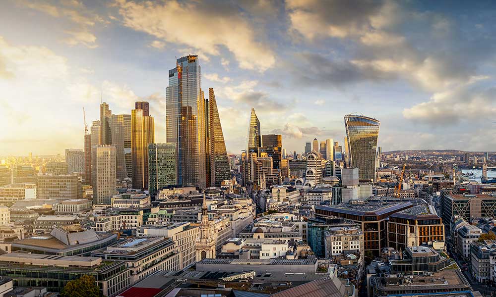 A panoramic view of a bustling cityscape at sunset, featuring numerous modern skyscrapers and buildings with diverse architectural styles, highlights the fruits of recent construction and investment plans. The partly cloudy sky, tinged with a golden hue from the setting sun, casts a warm light over the urban landscape.