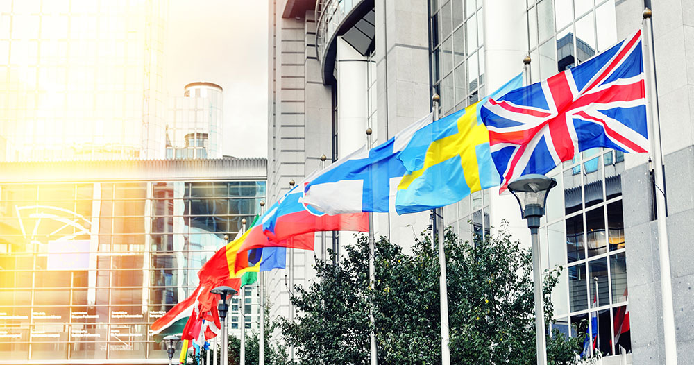 Various national flags, including those of the United Kingdom, Sweden, and others, are prominently displayed on flagpoles outside a modern glass building under a bright sky, symbolizing EU Representation.