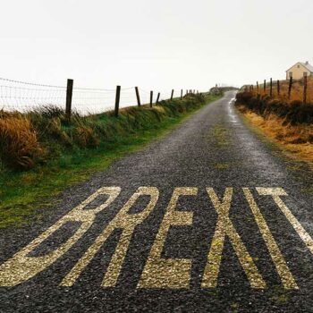 EU Representation a full guide for UK businesses 7 A narrow, rural road with "BREXIT" painted in large letters on the pavement. The road is flanked by grass and wire fences, leading towards a foggy horizon with a small white house visible in the distance on the right side. Amidst an overcast sky, "Welcome 2021! EU Representation" stands as a guide for UK businesses navigating forward.