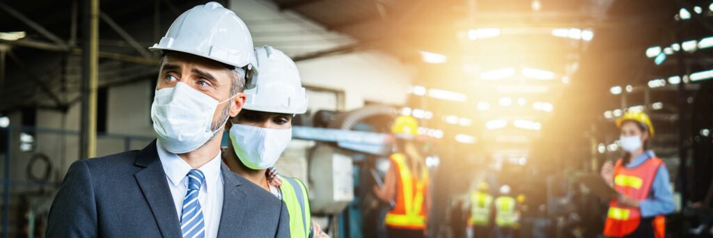 AdobeStock_372000533-web - JVR Consultancy Two individuals in safety helmets and face masks stand closely in a well-illuminated industrial setting. One wears a suit and tie, while the other wears a green jacket. Blurred workers in safety vests are visible in the background, showcasing their commitment to ISO 45001 certification standards.
