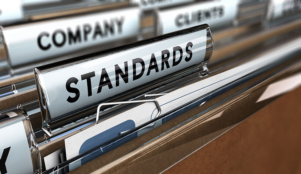 Close-up of file folders in a filing cabinet with labeled tabs. The prominently visible tab reads "STANDARDS," including important accreditations and ISO documents. Other partially visible tabs are "COMPANY" and "CLIENTS." The folders contain various documents and papers.