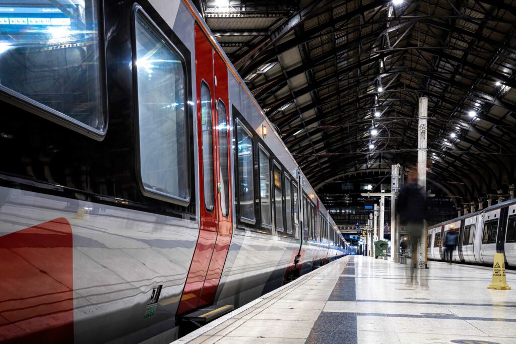 A long train is stationed on a well-lit platform inside a large, arched train station with a high ceiling and metal beams. The platform's tiles are shiny. One person is blurred in motion as they walk along the platform. Caution signs, placed near the train, adhere to strict Rail Licencing standards.
