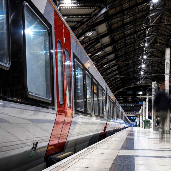 A long train is stationed on a well-lit platform inside a large, arched train station with a high ceiling and metal beams. The platform's tiles are shiny. One person is blurred in motion as they walk along the platform. Caution signs, placed near the train, adhere to strict Rail Licencing standards.
