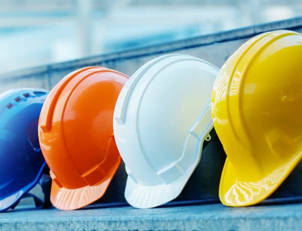 Four construction helmets are lined up on a set of concrete steps. From left to right, the helmets are blue, orange, white, and yellow. The background is slightly blurred, emphasizing the focus on the helmets—symbolizing the professionalism you'd expect when you become Constructionline Accredited.