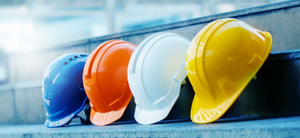 Four construction helmets are lined up on a set of concrete steps. From left to right, the helmets are blue, orange, white, and yellow. The background is slightly blurred, emphasizing the focus on the helmets—symbolizing the professionalism you'd expect when you become Constructionline Accredited.