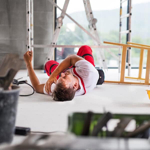 A construction worker lies on the floor, appearing to be in pain, with his arm clutching his chest. The scene includes a fallen ladder, scattered tools, and construction materials, indicating an accident on a worksite. A yellow hard hat is also on the floor nearby. This highlights the crucial need to become SSIP Accredited for safety compliance.