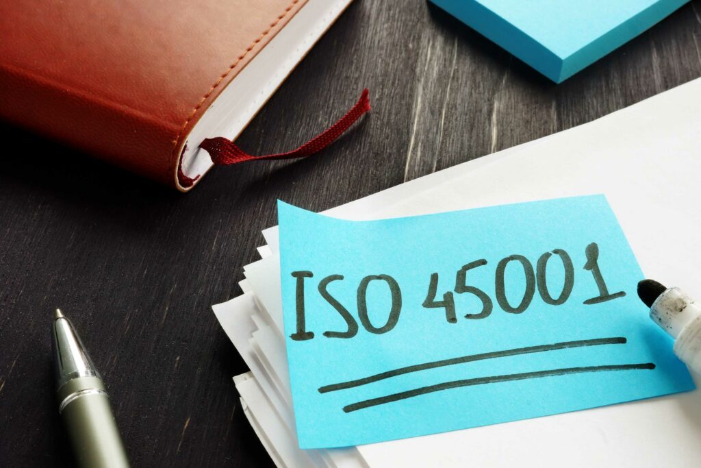 A blue sticky note with "ISO 45001" written in black marker is placed on a stack of white papers. Nearby, there is an open brown notebook with a red ribbon bookmark, a silver pen, and a black desk surface in the background, hinting at efforts to become ISO certified.