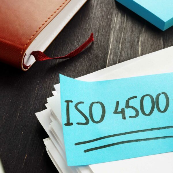 A blue sticky note with "ISO 45001" written in black marker is placed on a stack of white papers. Nearby, there is an open brown notebook with a red ribbon bookmark, a silver pen, and a black desk surface in the background, hinting at efforts to become ISO certified.
