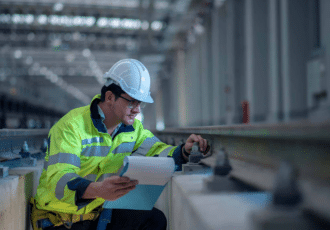 What is a Network Rail Principal Contractor Licence (PCL)? 1 A construction worker, wearing a hard hat and high-visibility jacket, examines rail tracks inside an industrial building. As one of the principal contractors, he holds documents or plans in one hand while using a tool to check the rail alignment or measurements with the other hand.