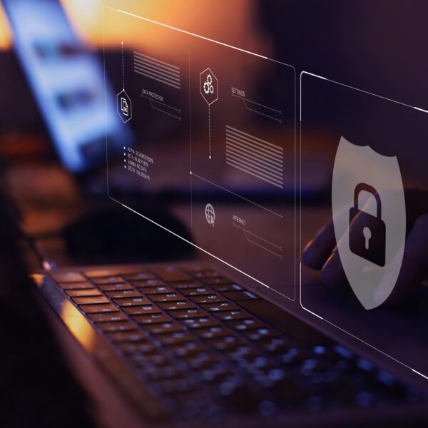 A dimly lit desk features an open laptop displaying digital security icons, including a shield with a lock symbol, as a person emphasizes cybersecurity. This setup sets the stage to become SSIP accredited, enhancing your workspace's commitment to top-notch security practices.